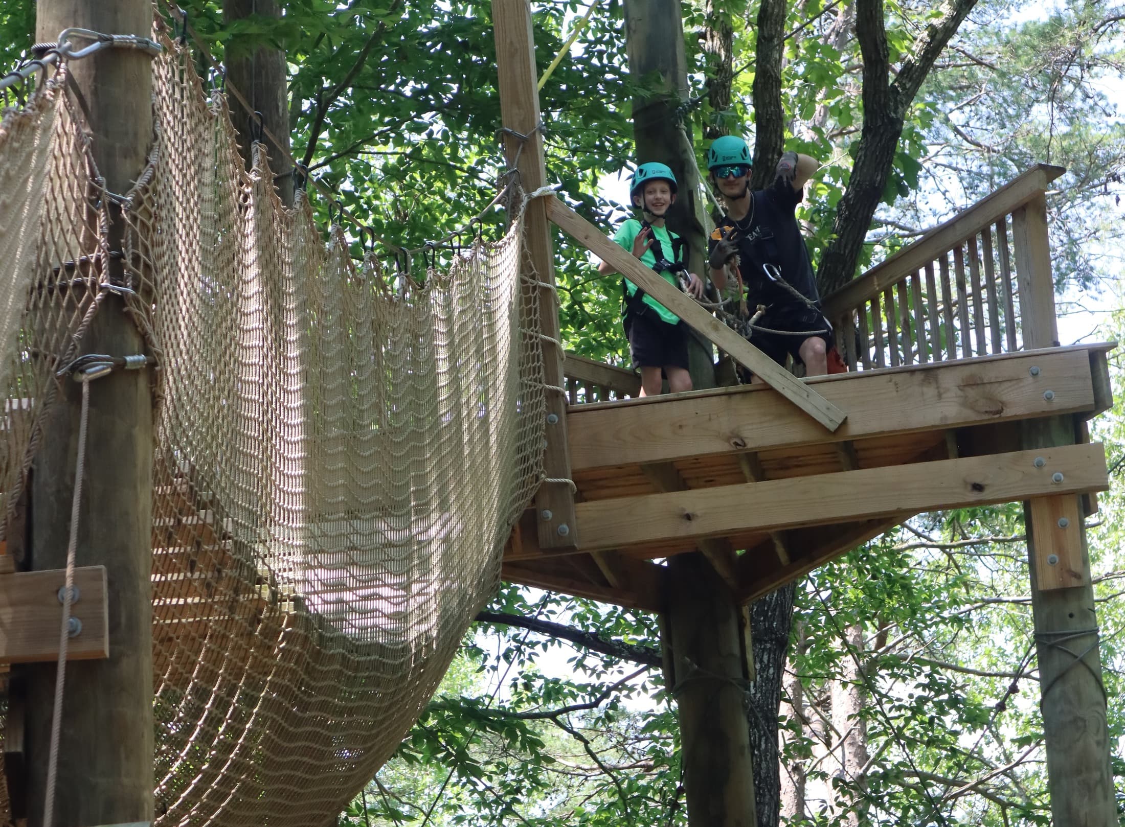 Two guests in helmets and harnesses on a treetop platform beside a rope net bridge on the aerial adventure course at Adventureworks Nashville West
